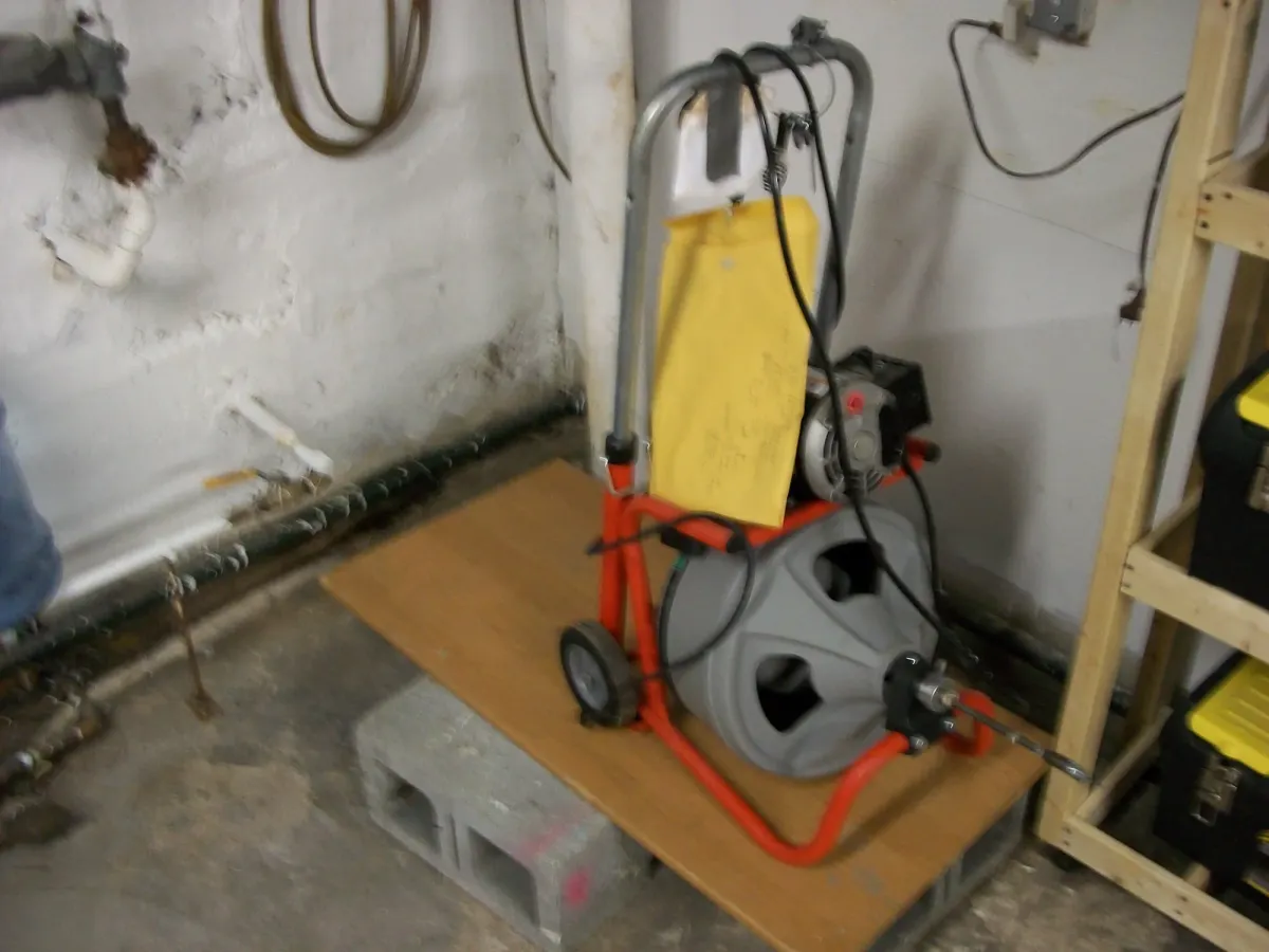 A drum-type drain cleaning machine set up beside a basement wall, its cable ready to feed into the drain