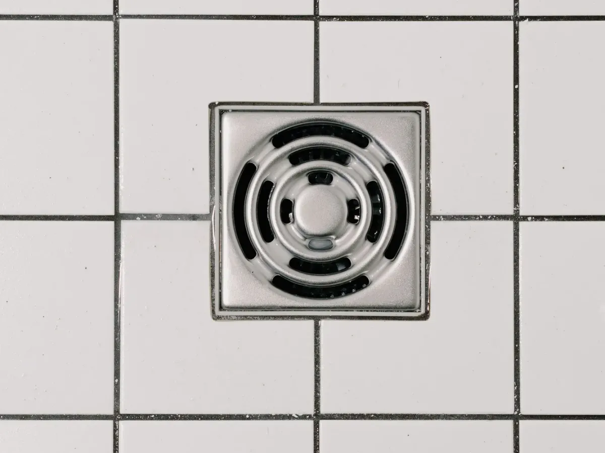 A square stainless steel floor drain set into a white tiled floor