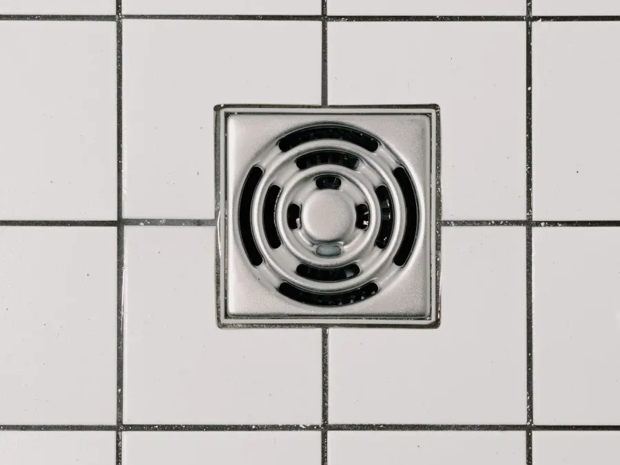A square stainless steel floor drain set into a white tiled floor